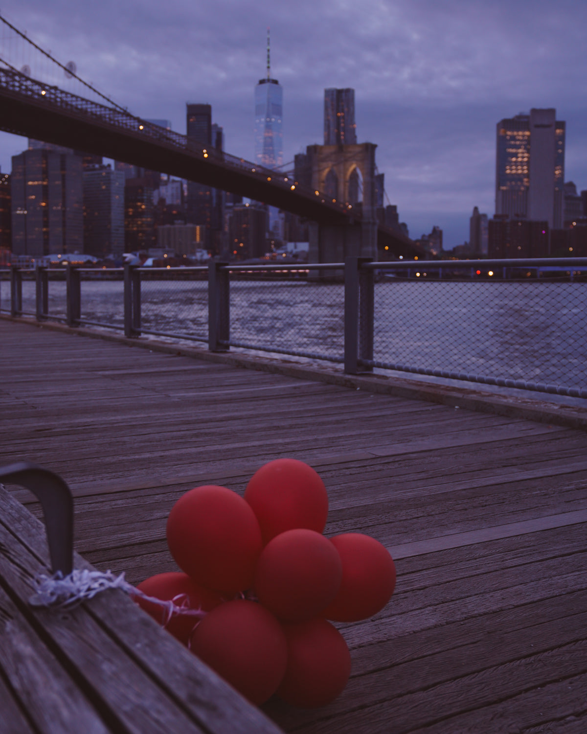 Red balloons falling over the side of a bench in front of the Brooklyn Bridge during a blue hour sunset