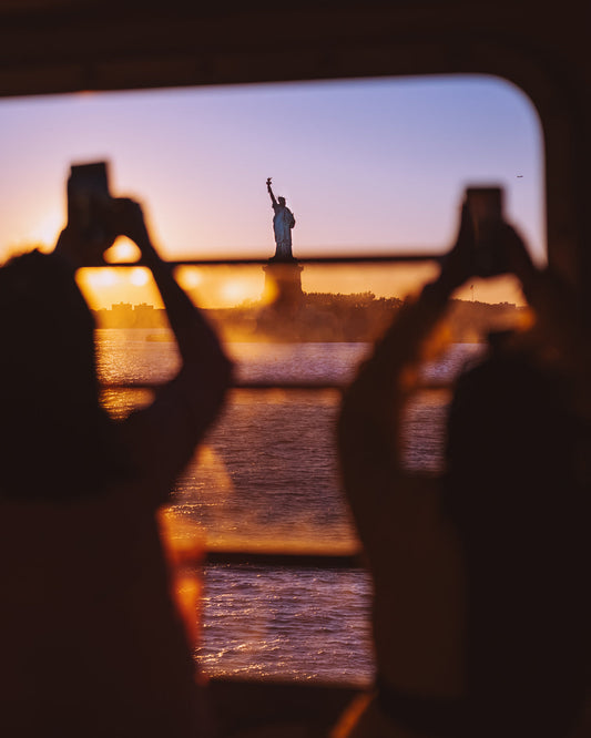 Two silhouettes taking photos on their phones of the Statue of Liberty through a ferry window at sunset