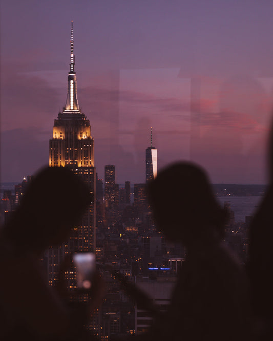 Two silhouettes looking at a phone sitting in a window sill looking out over the Empire State Building lit up