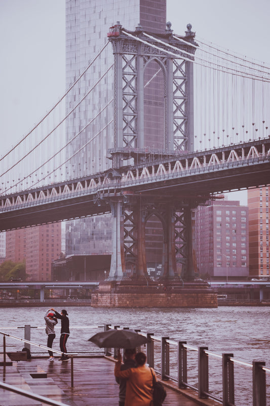 A couple under an umbrella watches a couple dancing in the rain in front of the Manahattan Bridge